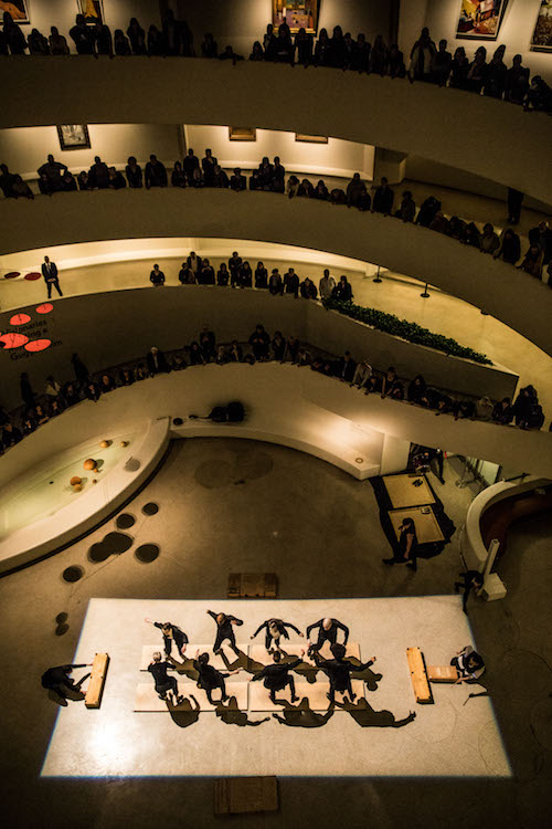A group of tap dancers tap on wooden boards as audience members peer down  to watch from above on the Guggenheim's various levels
